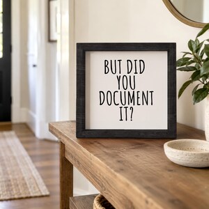 Black-framed documentation quote sign displayed on a wooden console beside a potted plant and shallow bowl.
