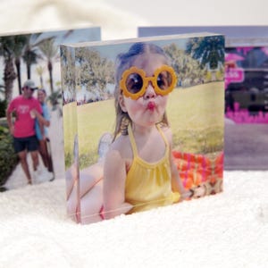 May include: A thick, clear acrylic block featuring a printed photograph of a child wearing yellow flower-shaped sunglasses and a yellow tank top, sitting on a colorful blanket outdoors in a park setting with trees and grass in the background.