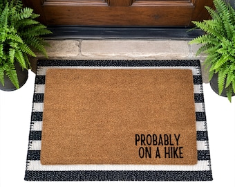 May include: A rectangular tan coir doormat layered over a black and white striped mat. The tan mat features the black text 'PROBABLY ON A HIKE' in the bottom right corner. The mats are positioned in front of a wooden door with two green ferns.