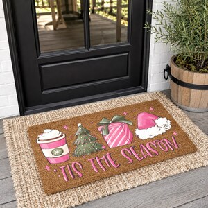 Op de afbeelding: Een rechthoekige bruine kokosmat met een roze koffiebeker, een groene kerstboom, een roze gestreept cadeau en een roze kerstmuts. De tekst 'TIS THE SEASON' staat in roze letters onderaan op de deurmat gedrukt.