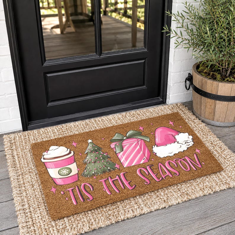 Op de afbeelding: Een rechthoekige bruine kokosmat met een roze koffiebeker, een groene kerstboom, een roze gestreept cadeau en een roze kerstmuts. De tekst 'TIS THE SEASON' staat in roze letters onderaan op de deurmat gedrukt.