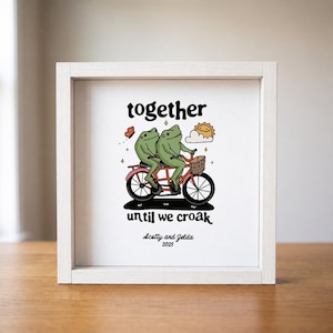 May include: A square white wooden frame containing a print of two green frogs riding a red tandem bicycle. Above the frogs is the text together and below is until we croak. Below that is the text Scotty and Zelda 2025 with a sun and butterfly.