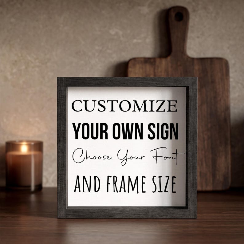 Black-framed personalized sign on a wooden kitchen counter in front of a cutting board, beside a lit candle, showing black lettering on a white insert.
