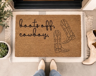 May include: A rectangular brown coir doormat with a white fabric border and fringe edges. The mat features black text that reads "boots off, cowboy." and a black line-art illustration of two western-style cowboy boots on the right side.