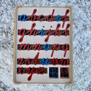 Może przedstawiać: Drewniana taca z literami alfabetu w stylu pisma odręcznego w kolorze czerwonym i niebieskim. Litery znajdują się w indywidualnych szczelinach i można je wyjmować. W małym przedziale pod tacą znajdują się dodatkowe litery.