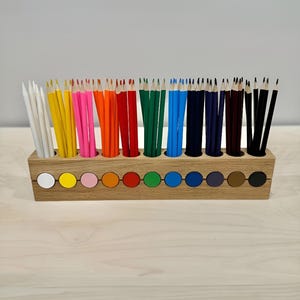 Puede incluir: Un porta lápices de madera con 12 compartimentos, cada uno con un círculo de color en la parte inferior. El porta lápices está lleno de lápices de colores, incluyendo blanco, amarillo, rosa, naranja, rojo, verde, azul, negro y marrón.