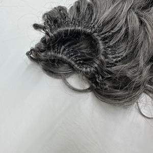 May include: A close-up of a dark gray curly hairpiece. The hairpiece is attached to a black mesh base with visible stitching. The hair is styled in loose, voluminous curls. The background is a plain white surface.