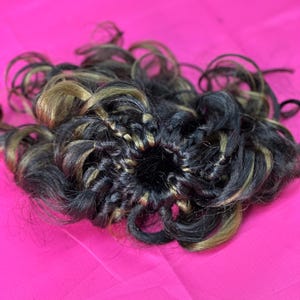 Puede incluir: Una pieza de cabello rizado, probablemente una peluca o extensión, de color marrón oscuro y rubio, se muestra sobre un fondo rosa brillante. El cabello está peinado con rizos apretados y parece sintético. La pieza de cabello está centrada.