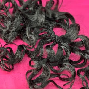 以下が含まれることがあります： 黒い巻き毛のエクステンションの束のクローズアップ。髪は自然なツヤがあり、ゆるく流れるカールにアレンジされています。背景は鮮やかなピンク色で、暗い髪と対照的です。