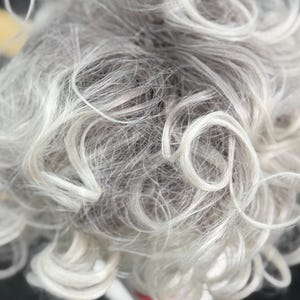 May include: A wig featuring short, curly hair in a blend of silver, white, and light gray tones. The hair is styled with loose, voluminous curls that create a textured appearance, with lighter white strands highlighting the top layers.