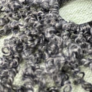 May include: A close-up of a dark grey curly hair extension. The hair is arranged in a circle, with the ends of the hair extending out from the centre.
