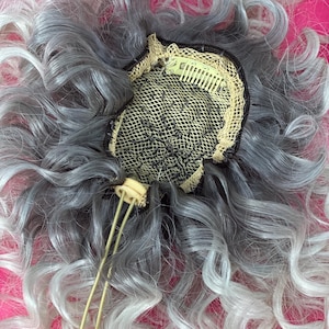 May include: A curly, gray and white wig with a mesh cap and hair clips. The wig has a mix of gray and white tones, with the curls being most prominent. The wig is set against a bright pink background.