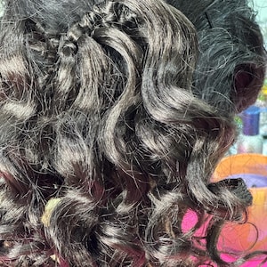 Puede incluir: Primer plano de cabello castaño oscuro y rizado. El cabello está peinado con una sección trenzada en la parte superior, que se transforma en rizos voluminosos y definidos. La textura parece suave y brillante, con reflejos naturales.