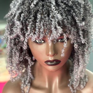 May include: A grey and silver curly wig with a side parting and bangs. The wig is made of synthetic hair and is styled in a short, layered cut.