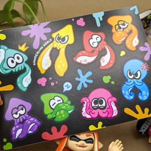 May include: A black background with colourful paint splatters and stickers of various coloured cartoon squids and octopuses. The stickers have different expressions and some have a crown on their head. The text "WANDERING WORLD" is written on the sticker sheet.