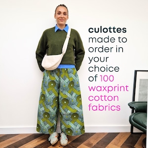 May include: A person wearing culottes made from waxprint cotton fabric. The culottes feature a green, yellow, and blue geometric pattern. The person is also wearing a green sweater, blue shirt, and a cream-coloured crossbody bag.