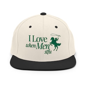 Op de afbeelding: Een witte en zwarte baseballpet met groene borduurwerk. Het borduurwerk zegt "I Love when Men stfu" en toont een cowboy die een paard berijdt.