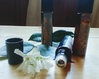 May include: Three glass roller bottles with black caps and floral designs. The bottles are on a wooden surface with white flowers and green leaves.