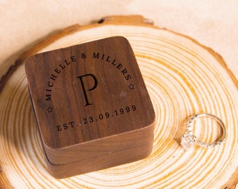 May include: A wooden ring box with a personalized inscription that reads "Michelle & Millers ☆ EST. 23.09.1999" and the letter "P" in the center. The box is sitting on a wooden slice.