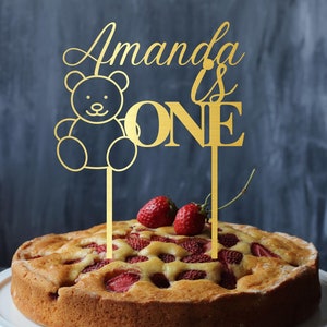 Könnte beinhalten: Goldener Tortendekor mit dem Text "Amanda ist eins" und einem Teddybär-Design. Der Dekor befindet sich auf einem Kuchen mit Erdbeeren.