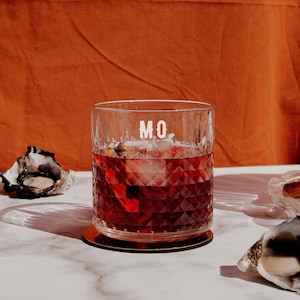 May include: A clear glass with a diamond pattern is filled with a dark red liquid and ice. The glass has the letters "MO" printed on the front. The glass is sitting on a gold coaster on a white marble surface.