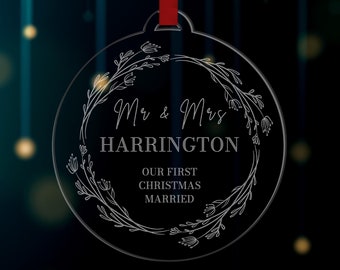 Pode incluir: Um ornamento redondo transparente com um design de coroa de flores branca. O ornamento tem o texto "Mr & Mrs HARRINGTON" e "OUR FIRST CHRISTMAS MARRIED" inscrito nele.