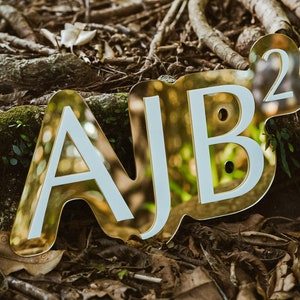 以下が含まれることがあります： 茶色と緑色の背景に「AJB²」と綴られた金色の文字。文字は立体で、白い輪郭があります。