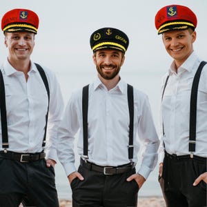 May include: Three men wearing white shirts, black suspenders, and black pants. Each man is wearing a captain's hat. Two hats are red with gold trim and the third is black with gold trim. The hats have text and an anchor emblem.