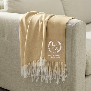 May include: A beige throw blanket with white fringing is draped over a sofa arm. The blanket has a white embroidered design with the initials "L/Z", a laurel wreath, and the names "Lars & Zariffi" with the date "14.09.2025".