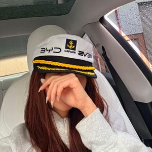 May include: White captain's hat with black and gold trim, featuring the text "343 CAPTAIN EV" and an anchor emblem. The hat is worn in a car. The hat has a black brim with gold rope trim.