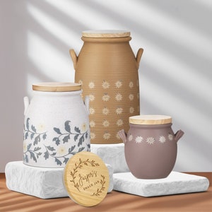 May include: Three ceramic jars with wooden lids and floral patterns. The jars are in different sizes and colors: brown, white, and purple. The largest jar has a floral pattern with a light brown background. The middle jar has a white background with a floral pattern in shades of blue and white. The smallest jar has a purple background with a white floral pattern. The lid of the smallest jar has a wooden circle with the text "Jasper's Snack Jar" in brown lettering.