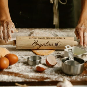May include: A wooden rolling pin with the text "Premium Quality From Mum's Kitchen" and the name "Brynlee" with two hearts. The rolling pin is on a wooden surface with flour and other baking supplies.