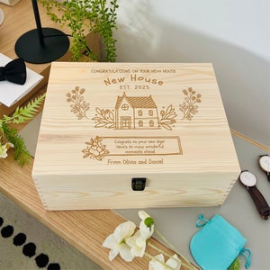 May include: A wooden box with a personalised message for a new home. The box is engraved with the words "Congratulations on your new house, New House Est. 2025."  The box also has a small house illustration with flowers and the message "Congrats on your new digs! Here's to many wonderful moments ahead. From Olivia and Daniel."