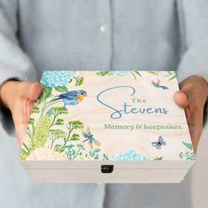 Puede incluir: Una caja de madera para recuerdos con un acabado de madera clara. La tapa presenta un diseño de acuarela de hortensias azules, hojas verdes, un pájaro azul, una libélula y una mariposa. El texto "The Stevens Memory & Keepsakes" está impreso en la tapa.