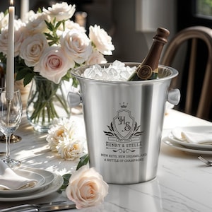 May include: A silver champagne bucket with a personalized inscription that reads "Est. 2023 Henry & Stella New Keys, New Dreams, and a Bottle of Champagne". The bucket is filled with ice and a bottle of champagne.
