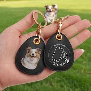 May include: Two black leather keychains with gold-colored hardware. One keychain features a smiling dog portrait, the other has a phone icon, paw print, and the name "SIMBA". The text "TAP YOUR PHONE" is also visible.
