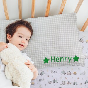 May include: A gray and white gingham pillow with green stars and the name "Henry" embroidered on it. The pillow is in a crib with a white and green sheet with a farm animal print.
