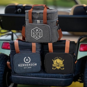May include: Three insulated golf cooler bags in grey, navy, and black, with brown handles and straps. Each bag has a front pocket with a golf-themed logo and text. The bags are stacked on a golf cart.