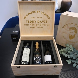 以下が含まれることがあります： 「Brothers by Chance, Est. 2024, Teddy Davis, Friends by Choice, Love, Louis」と刻まれた、蝶番付きの蓋が付いた木製の箱。箱の中にはワインが3本入っていて、そのうちの1本は「2009」と金色のラベルが貼られたシャンパンボトルです。