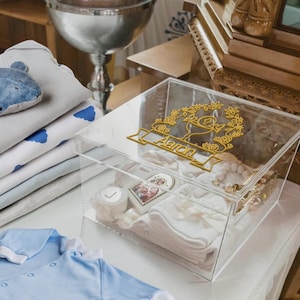 May include: A clear acrylic box with a gold floral design and the name "Aaron" on the lid. Inside the box are white baby clothes, a small religious icon, and a white candle. Baby clothes and a whale-shaped toy are visible nearby.