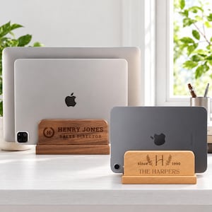 May include: Two wooden device stands holding electronic tablets and laptops. The darker wood stand is engraved with "HENRY JONES SALES DIRECTOR" and a circular logo. The lighter wood stand is engraved with "since 1990", "H", and "THE HARPERS".