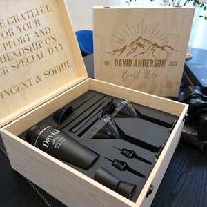 Puede incluir: Una caja de madera con una inscripción grabada en negro "David Anderson Best Man" y un diseño de cordillera. La caja contiene una coctelera negra, dos copas de martini, un colador de cóctel, un jigger y un tapón de botella.