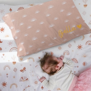 May include: A pink pillowcase with a white sun pattern and the name "Bella" embroidered in yellow. The pillowcase is on a white sheet with a similar sun pattern in brown, orange, and yellow.