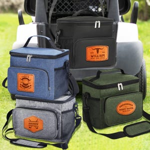 May include: Several insulated lunch bags in shades of blue, black, grey, and green. Each bag has a personalised leatherette patch with text and graphics. They feature a top handle, shoulder strap, and a side mesh pocket.
