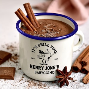 May include: White enamel mug with a blue rim filled with chocolate milk and topped with chocolate shavings. The mug has a black and white logo that says "It's Chill Time" with a cartoon character holding a mug and the text "Henry Jones's Babyccino" below.