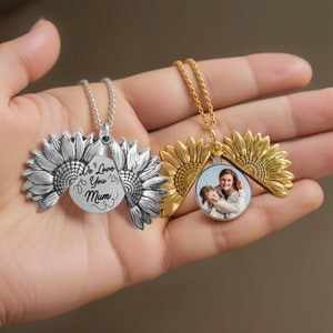 Könnte beinhalten: Zwei Sonnenblumen-Medaillon-Halsketten, eine in Silber und die andere in Gold. Ein Medaillon enthält ein Foto einer Mutter und ihrer Tochter, und das andere Medaillon trägt die Inschrift "We Love You Mum" auf der Vorderseite.