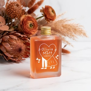 May include: A rectangular frosted glass bottle filled with amber liquid, featuring a gold screw cap. The front displays a white line drawing of two figures holding a heart with the text Olivia + Matt and the date 4.08.2026 printed below.