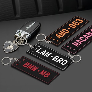 Op de afbeelding: Een set van vijf zwarte sleutelhangers met verschillende automerklogo's en namen. De sleutelhangers zijn gemaakt van zwart rubber met witte letters. De logo's omvatten Lamborghini, AMG, Lam Bro, BMW en Macan.