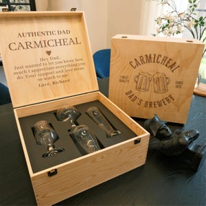 May include: A wooden box with a personalized message for Dad. The box contains four glass beer mugs and a small wooden figurine. The box is labeled "Authentic Dad Carmichael" and "Dad's Brewery".
