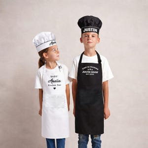 May include: Two children in personalized chef attire. One wears a white chef's hat and apron with "Amelia" and "Baby Kitchen Star". The other wears a black chef's hat and apron with "Justin" and "Chef in Training".
